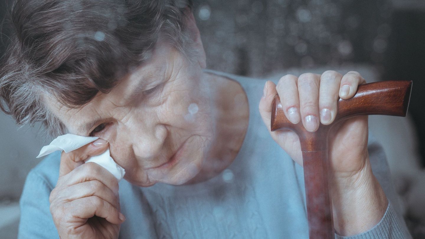 Alzheimer’s disease: woman with walking stick looking down and holding a handkerchief to her eye.