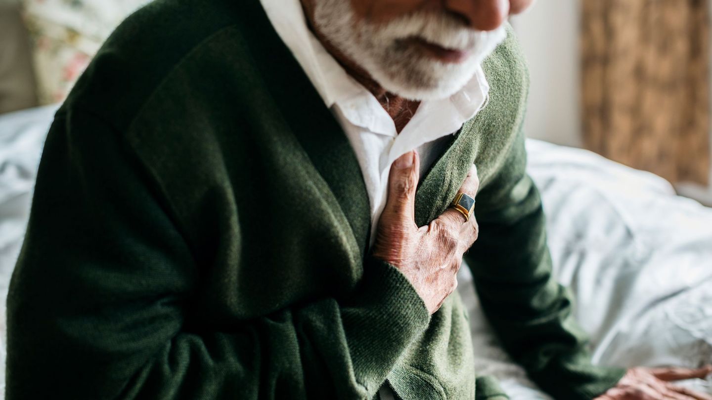 Angina pectoris: old man sitting on a bed looking tired and clutching his chest.