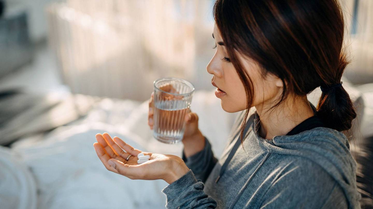 Eine junge Frau in häuslicher Umgebung betrachtet einige Tabletten, die auf ihrer linken Handfläche liegen. In der rechten Hand hält sie ein Wasserglas.
