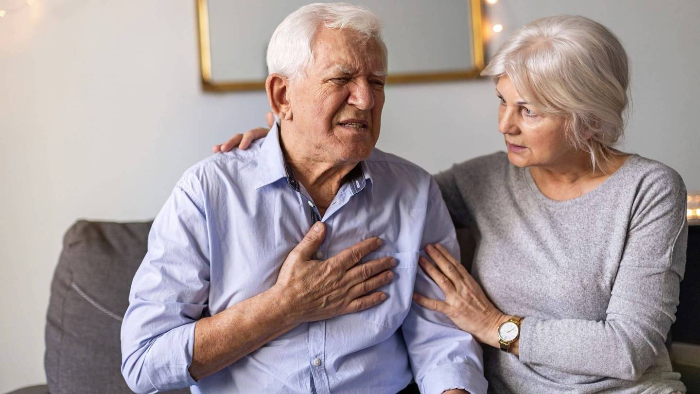 Older man sitting on a sofa with his hand on his chest grimacing in pain. An older woman is sitting next to him and comforting him with her hands on his shoulder and arm, looking at him and is clearly worried.