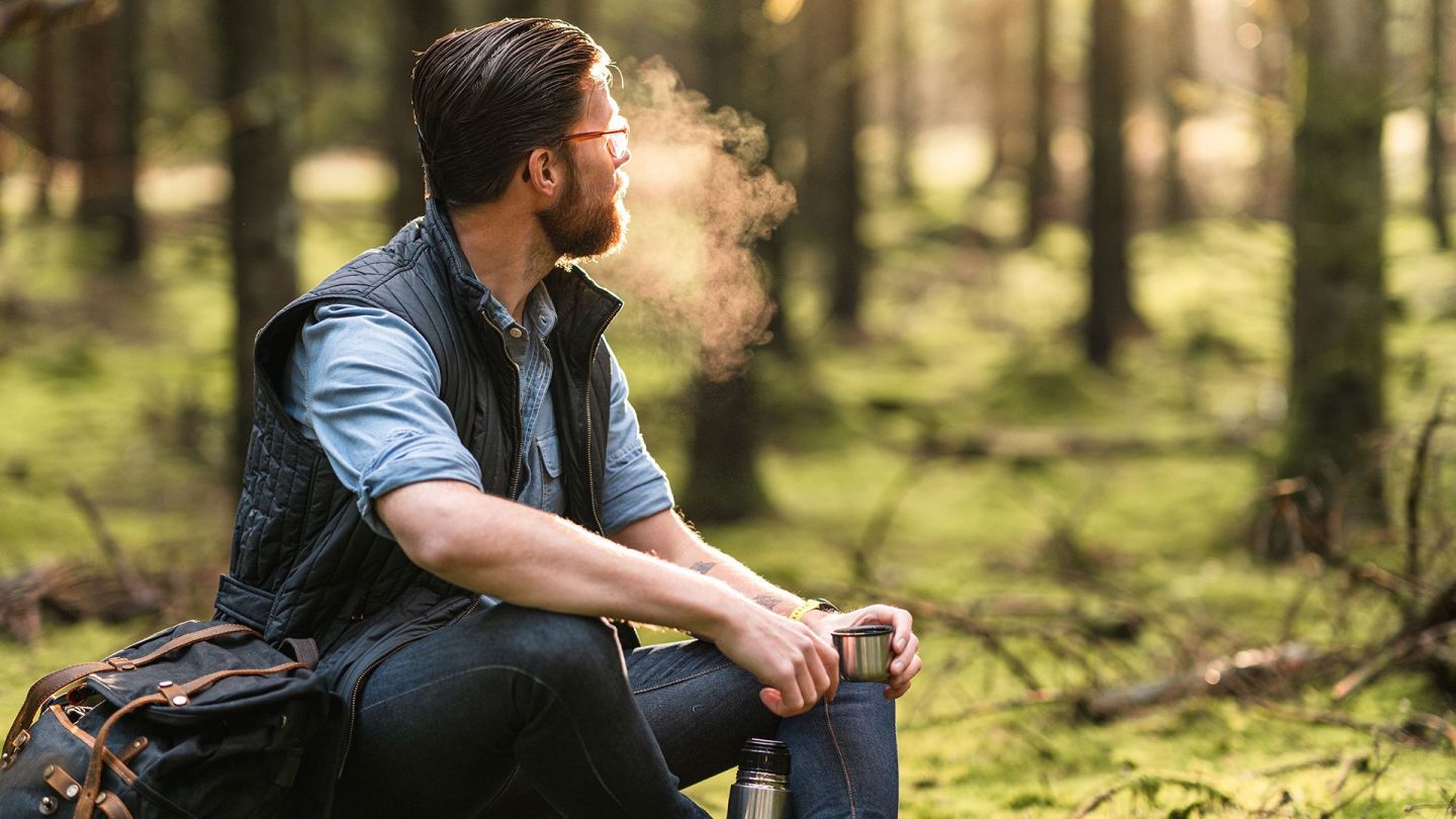 Ein Mann sitzt in einem Wald auf einem Koffer und hält den Becher einer Thermoskanne in einer Hand. Er blickt in die Ferne, zwischen Bäumen hindurch - vor seinem Gesicht verdunstet eine Atemwolke.