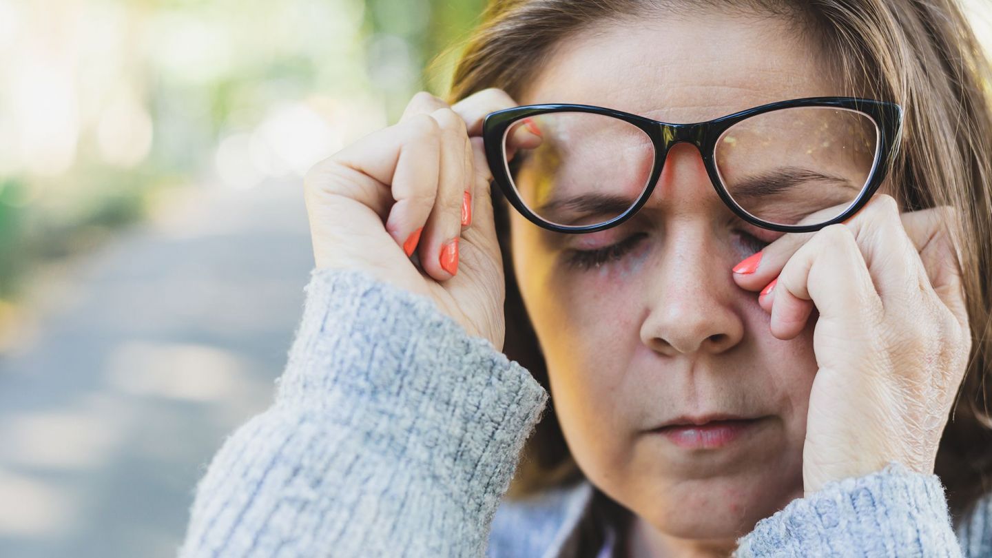 Blepharitis: women wearing glasses pushing her glasses up slightly with one hand and touching her closed eye with the other.