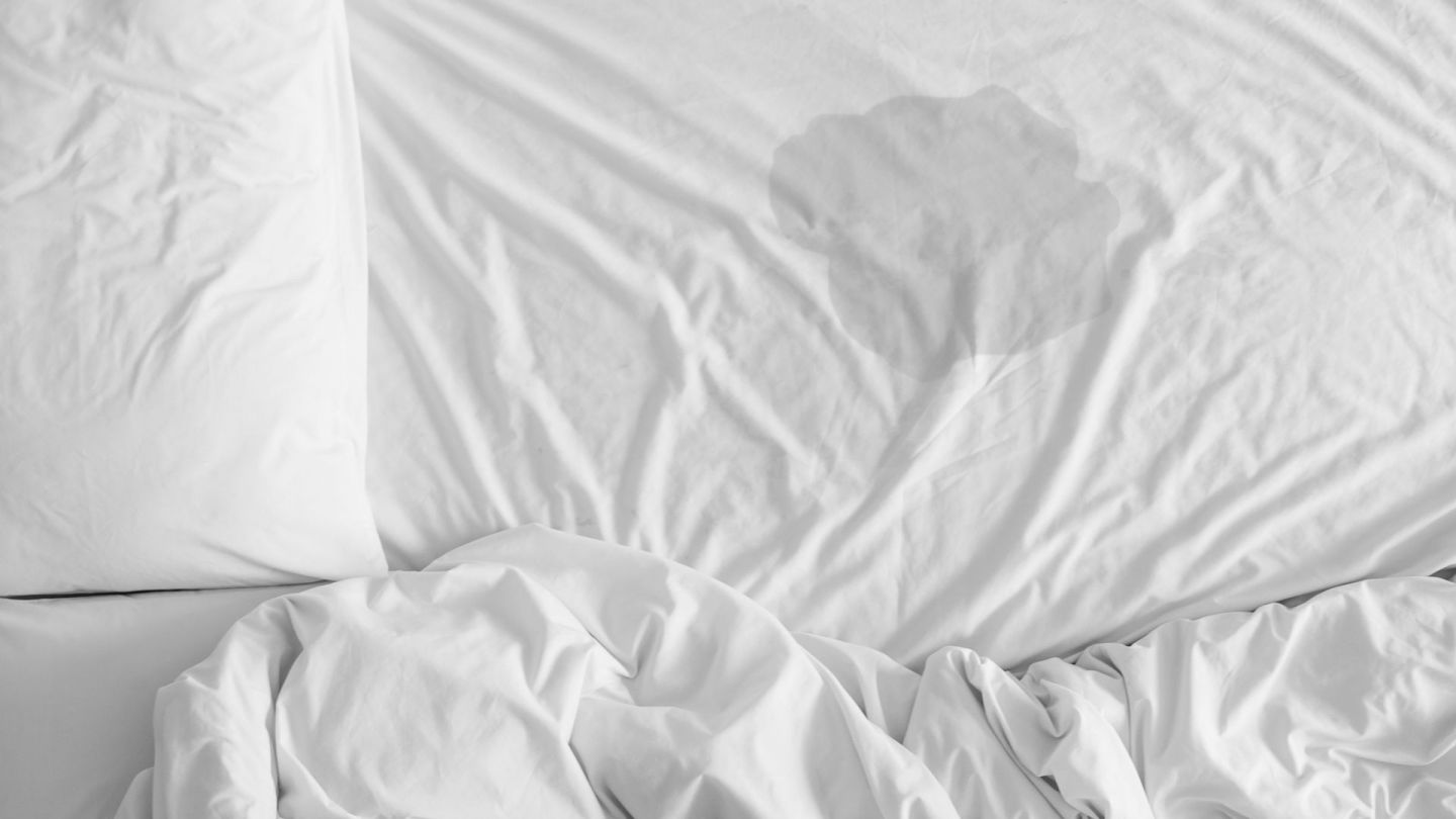 Bettnässen: In der Mitte eines weißen Bettbezugs, der über eine Matratze gezogen ist, ist ein größer Fleck zu sehen, der von einer Flüssigkeit stammt, offensichtlich Urin.