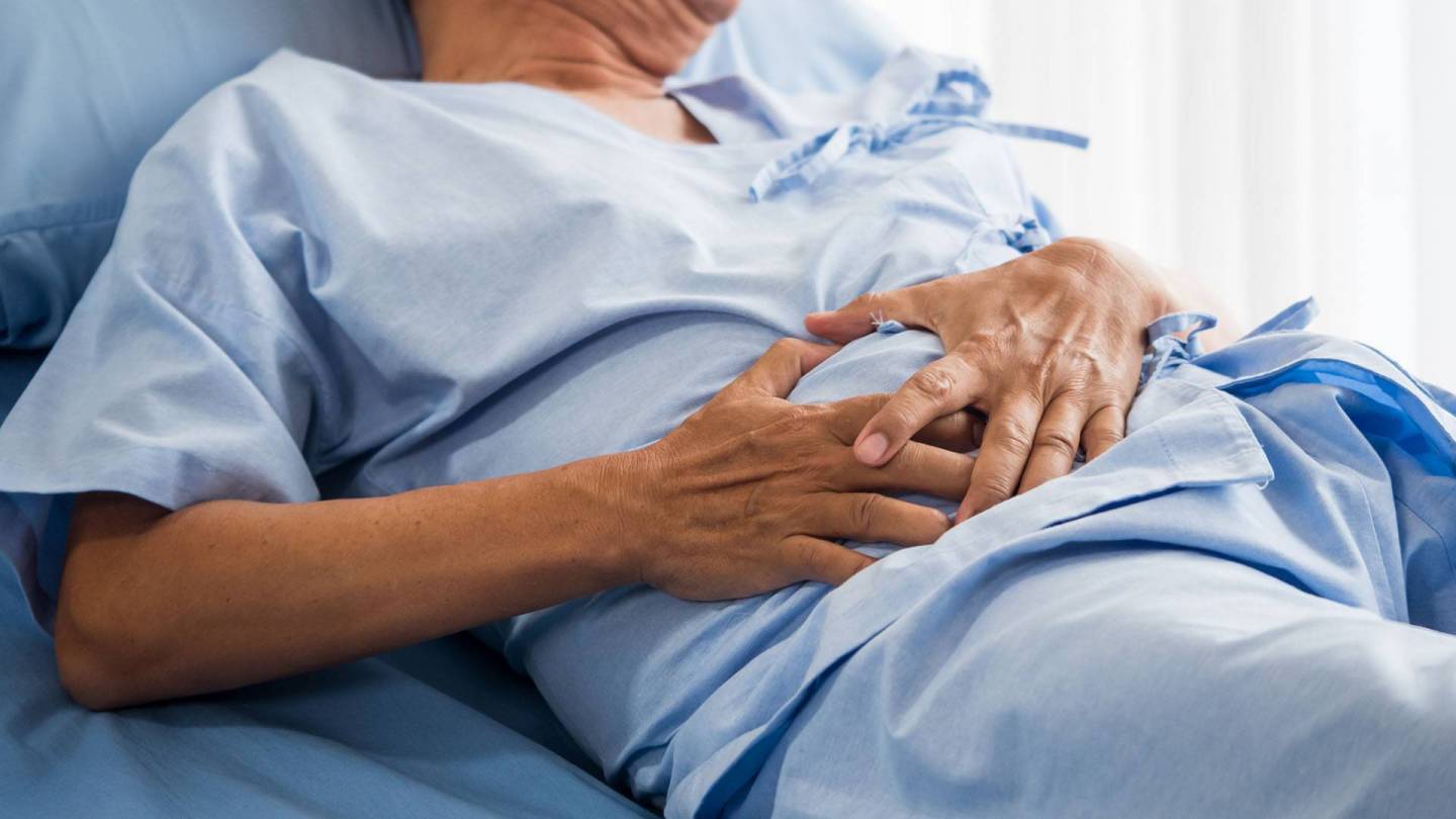 Ein Patient liegt mit medizinischer Kleidung auf einem Intensivbett und hält sich den Bauch.