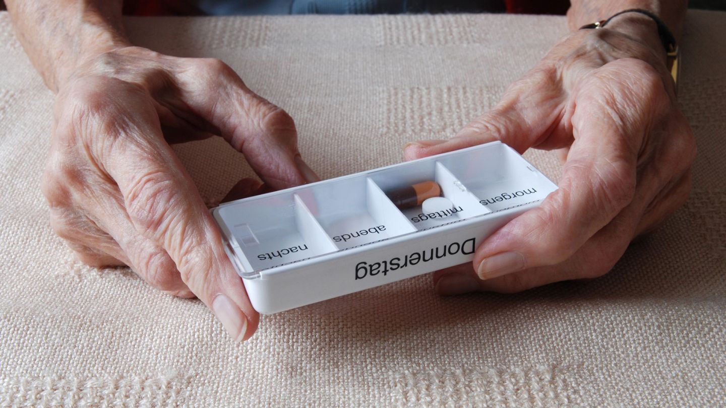 Electronic medication plan (eMP): older woman holding a plastic dosette box. The box is labeled “Thursday”. One of the compartments contains an oval tablet and a capsule.