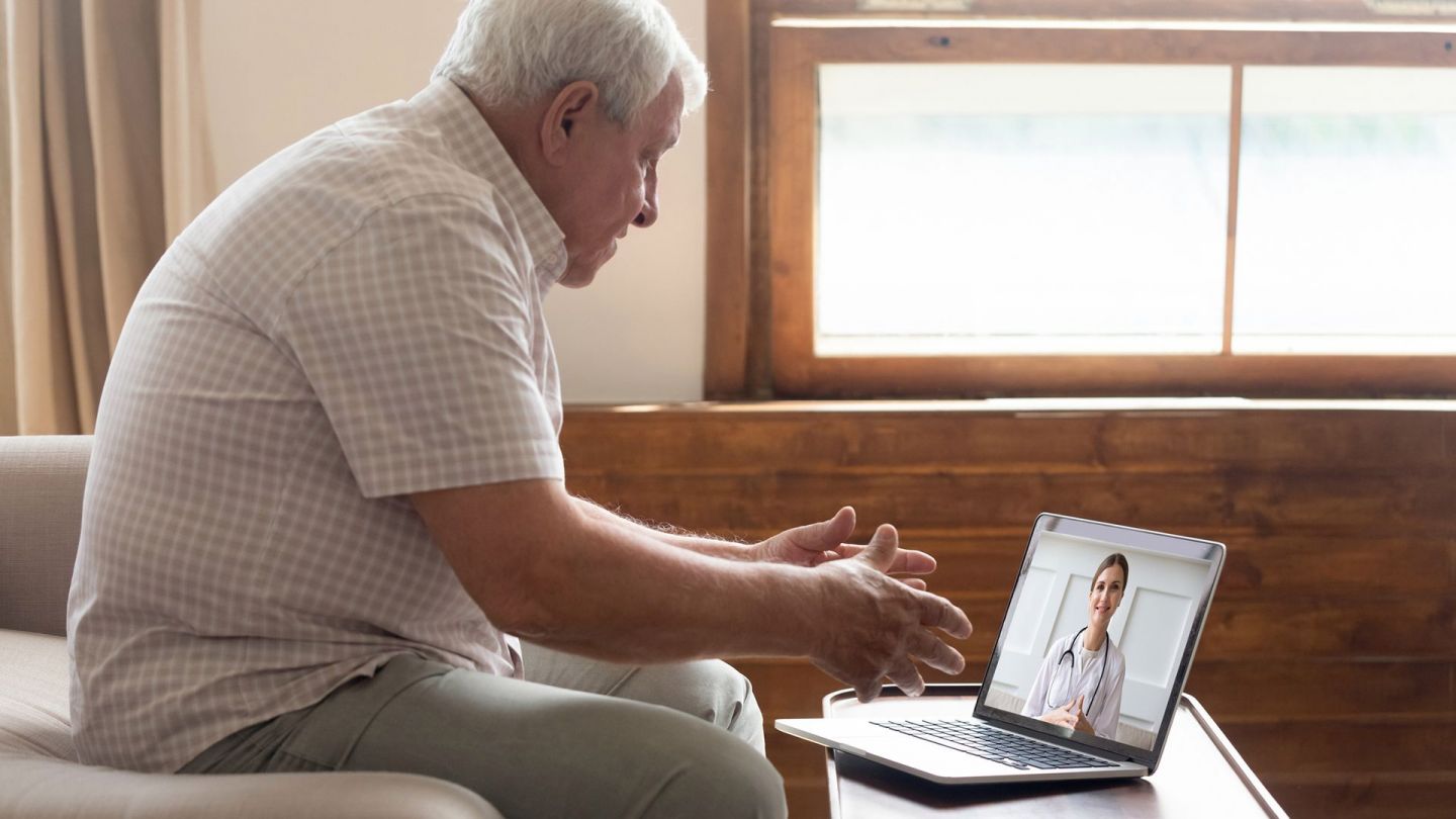 Online video appointment: older man sitting in front of a laptop which is standing on a table. On the screen he sees a younger woman in a white coat looking at the man.