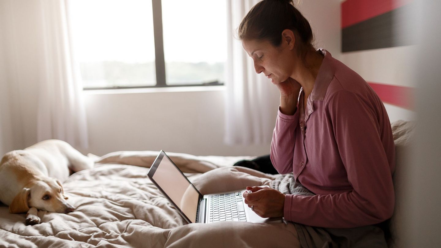 A woman sits on her bed with her laptop open.