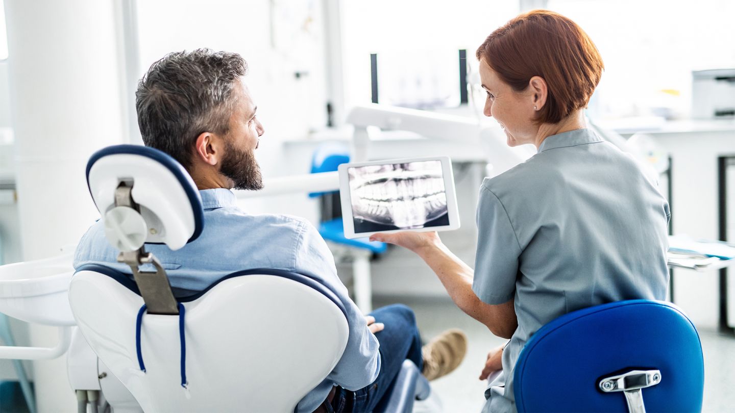 E-Zahnbonusheft: Ein Patient sitzt auf dem Behandlungstuhl beim Zahnarzt. Die Ärztin sitzt neben ihm und zeigt ihm die Röntgenaufnahme seines Gebisses auf einem Tablet.