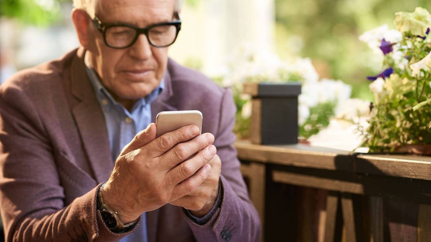 Older man in glasses sitting outside a cafe looking at his smartphone screen.