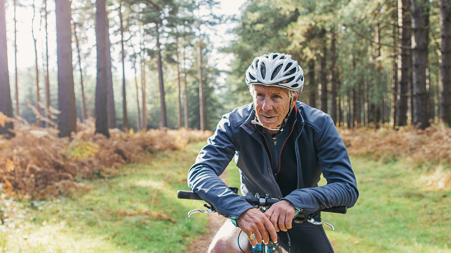 Ernährung und Bewegung im Alter: Ein älterer Mann in Fahrradkleidung pausiert während einer Radtour im Wald und stützt sich dabei auf den Lenker seines Fahrrads.