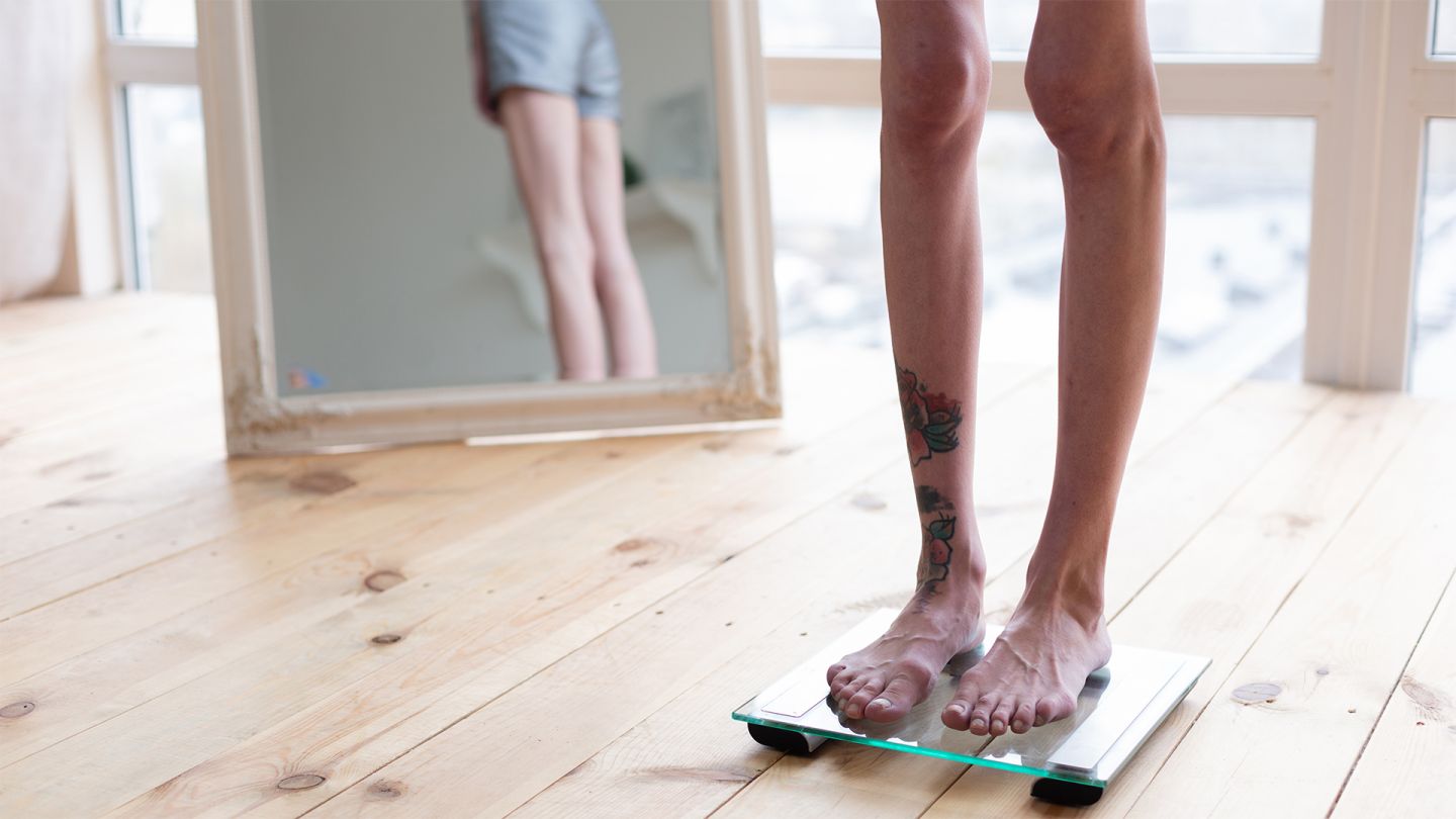 اضطراب التغذية: امرأة تعاني من نقص الوزن تقف على ميزان، وظهرها نحو مرآة تعكس كامل الجسم.