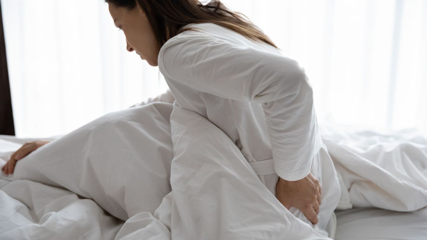 Fibromyalgie: Eine Frau sitzt leicht nach vorne gebeugt in einem Bett. Mit einer Hand fasst sie sich an den Rücken.