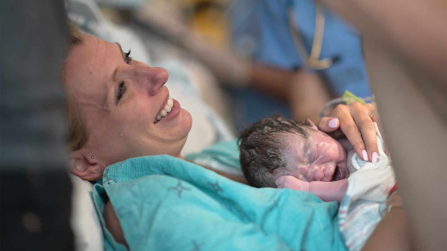 Childbirth: woman lying in a hospital bed holding her newborn baby in her arms. She looks happy and exhausted.