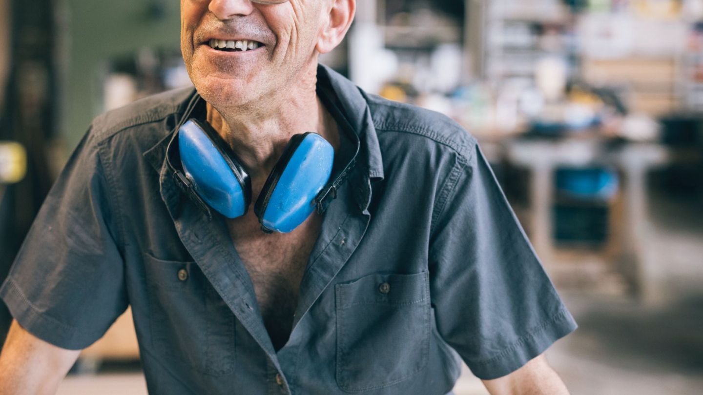 Ein älterer Mann trägt Arbeitskleidung, unter anderem einen Hörschutz. Der Mann lächelt.