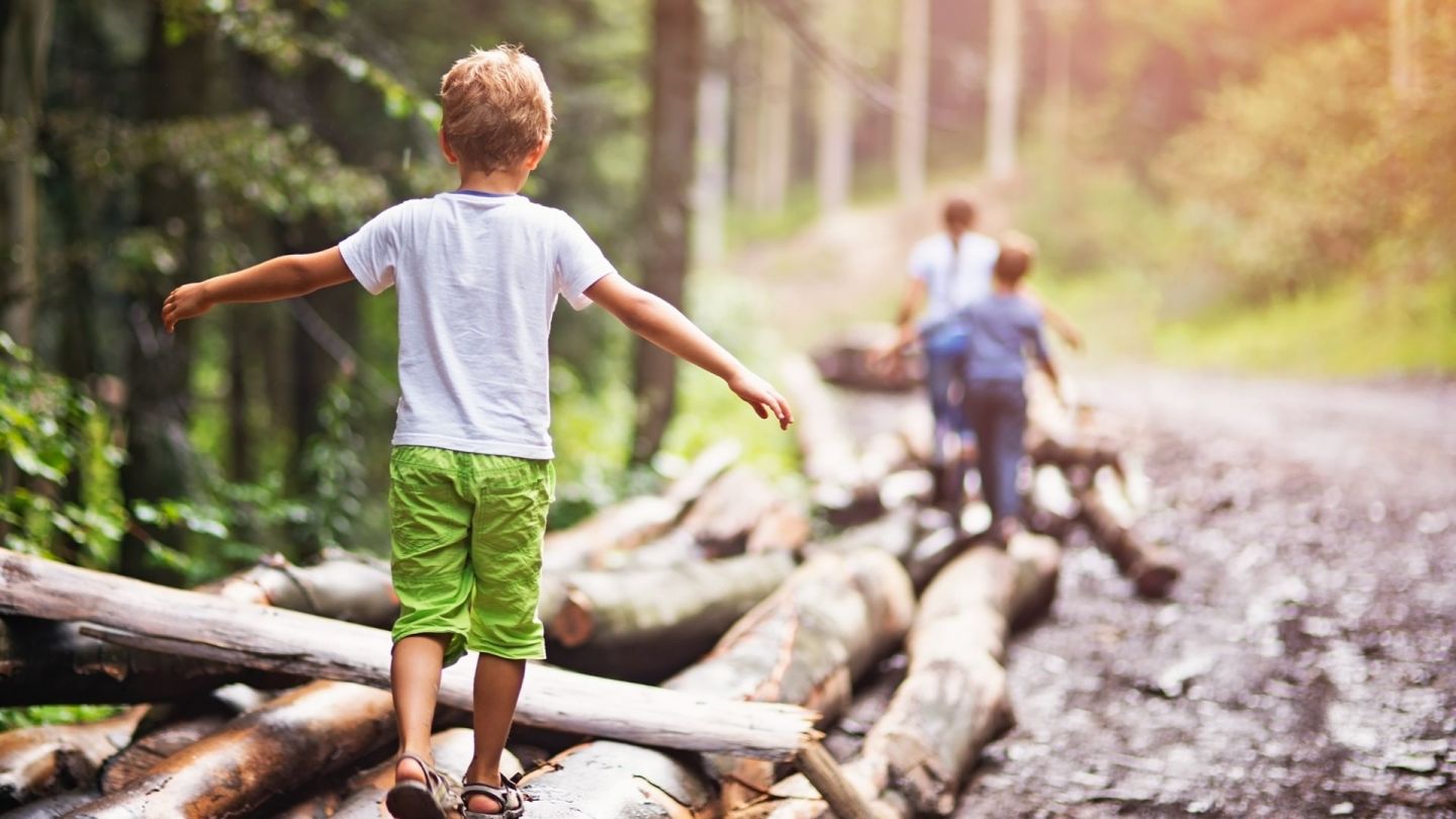 Kinder spielen an einem Waldweg, ein Junge balanciert mit ausgebreiteten Armen auf einem Baumstamm.