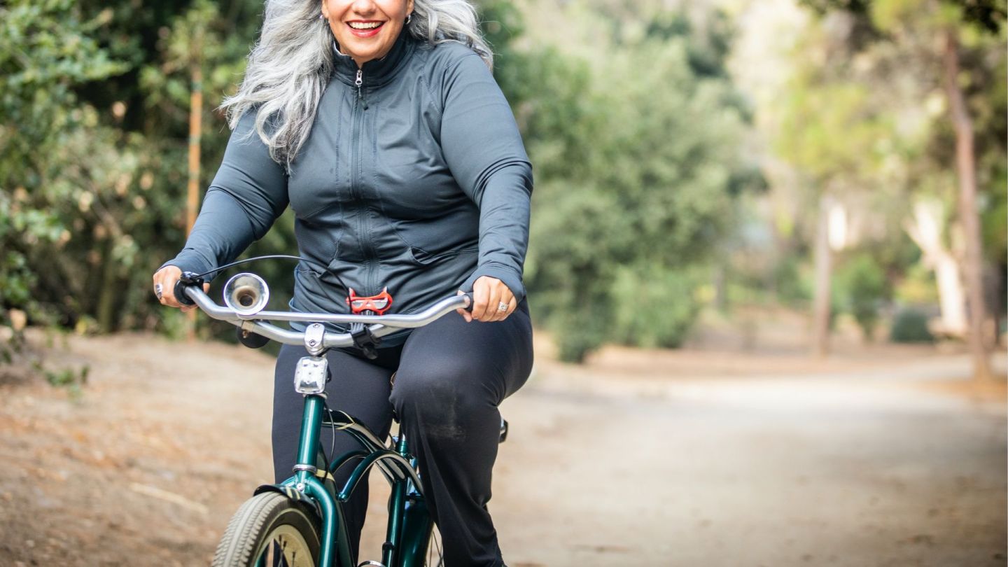 Eine ältere Frau fährt mit einem Fahrrad einen Waldweg entlang. Sie lächelt.