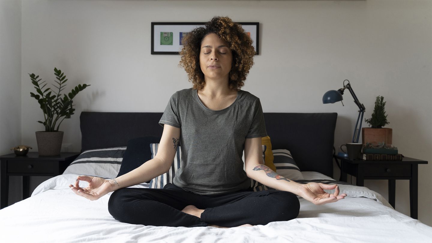 Rahatlamış ve sağlıklı: Bir kadın yatağında bağdaş kurup oturuyor ve meditasyon yapıyor. İki eli de dizlerinin üzerinde ve elleri chinmudra pozisyonunda. Gözlerini kapatmış ve nefes veriyor.