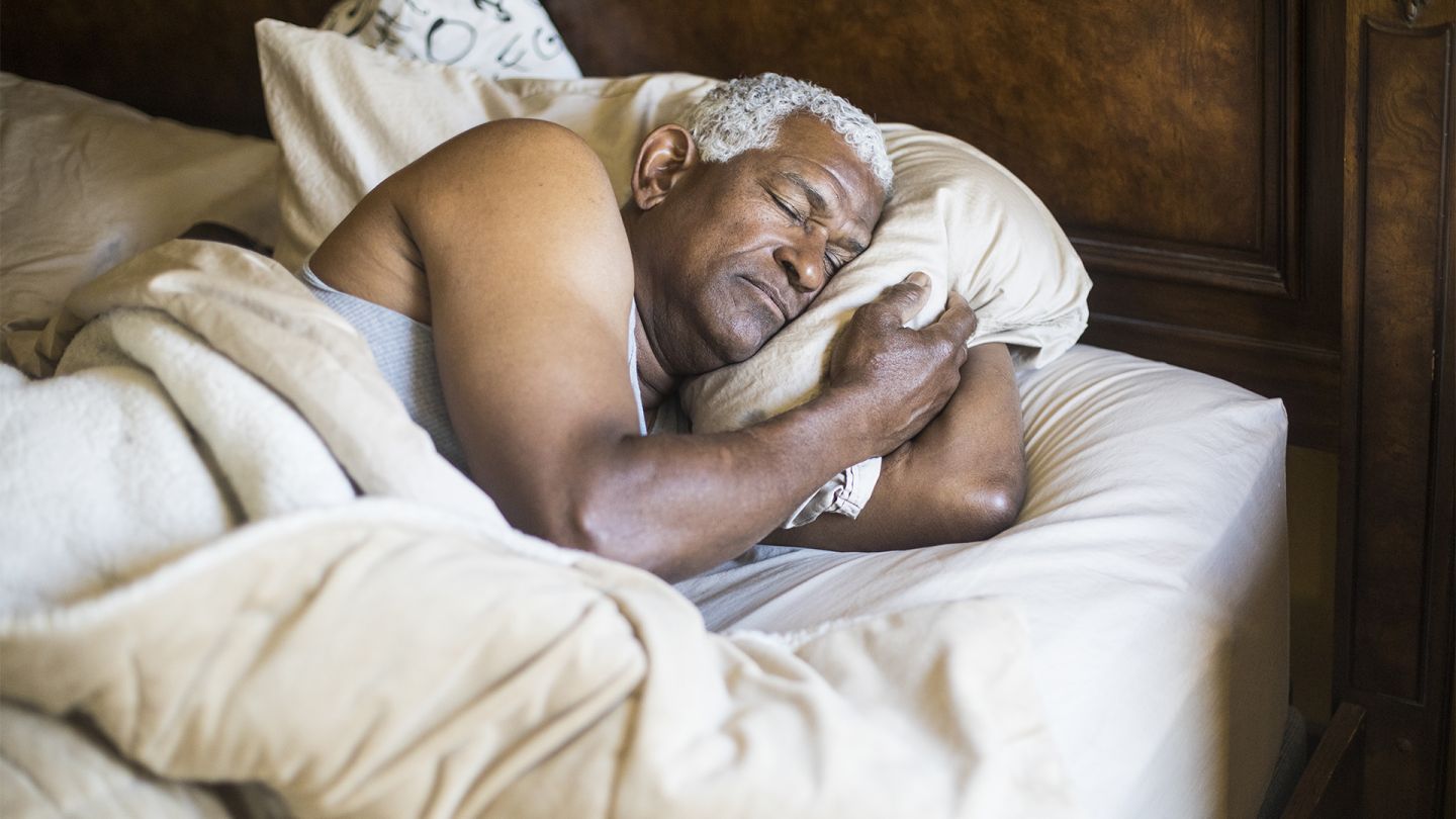 Healthy sleeping in later life: older man lying on his side in bed sleeping. He has his arm bent, clutching his pillow between his head and his arm.