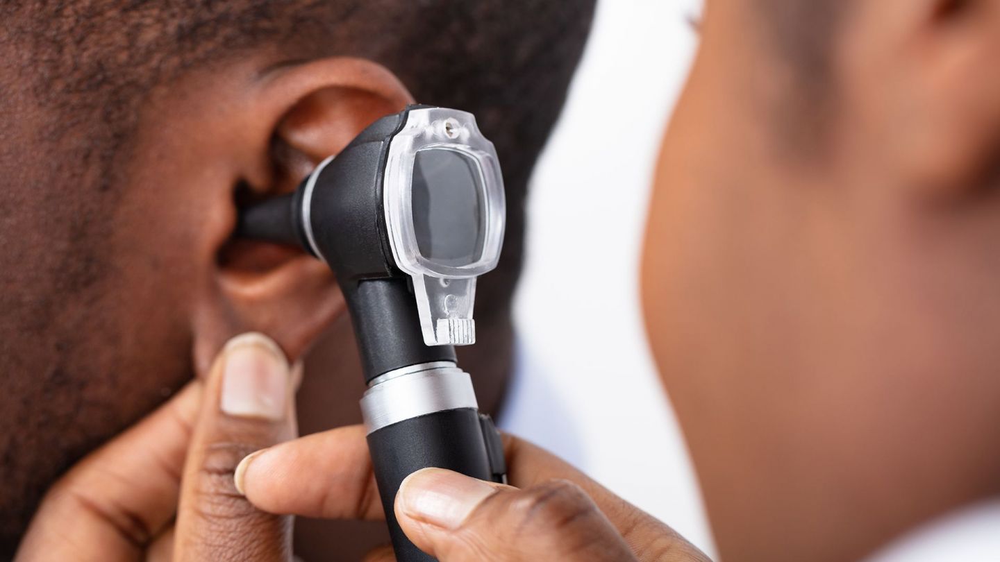 Ein Hals-Nasen-Ohren-Arzt hält mit einer Hand das Ohrläppchen eines Mannes und schaut mit einem Otoskop in das Ohr.