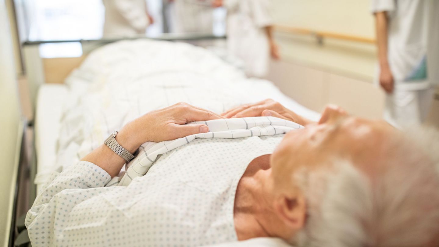 Ein älterer Mann liegt mit dem Rücken auf einem Behandlungsbett, das durch ein Krankenhauses geschoben wird. 