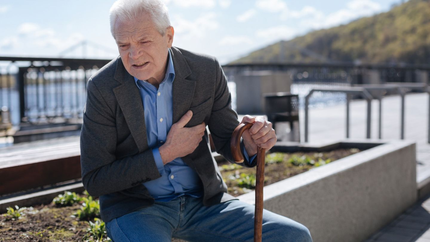Heart failure: older man sitting on a rock holding his heart with one hand. His face is tense and he is grimacing with pain.