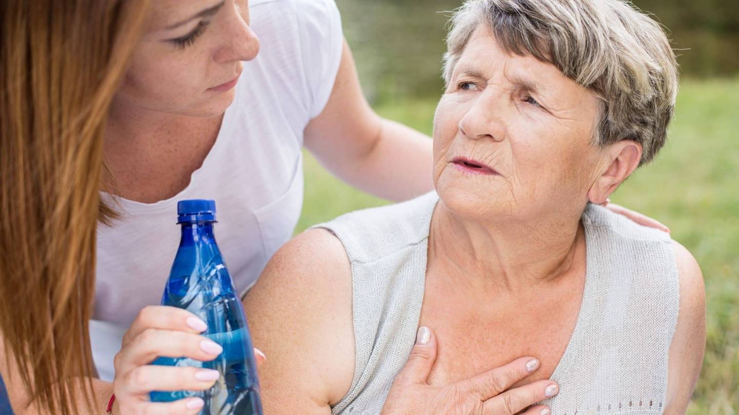 Eine ältere Frau im Sommershirt hat die Hand auf die Brust gelegt. Sie wirkt angestrengt und dreht sich zu einer jüngeren Frau um, die eine Flasche Wasser in der Hand hält.