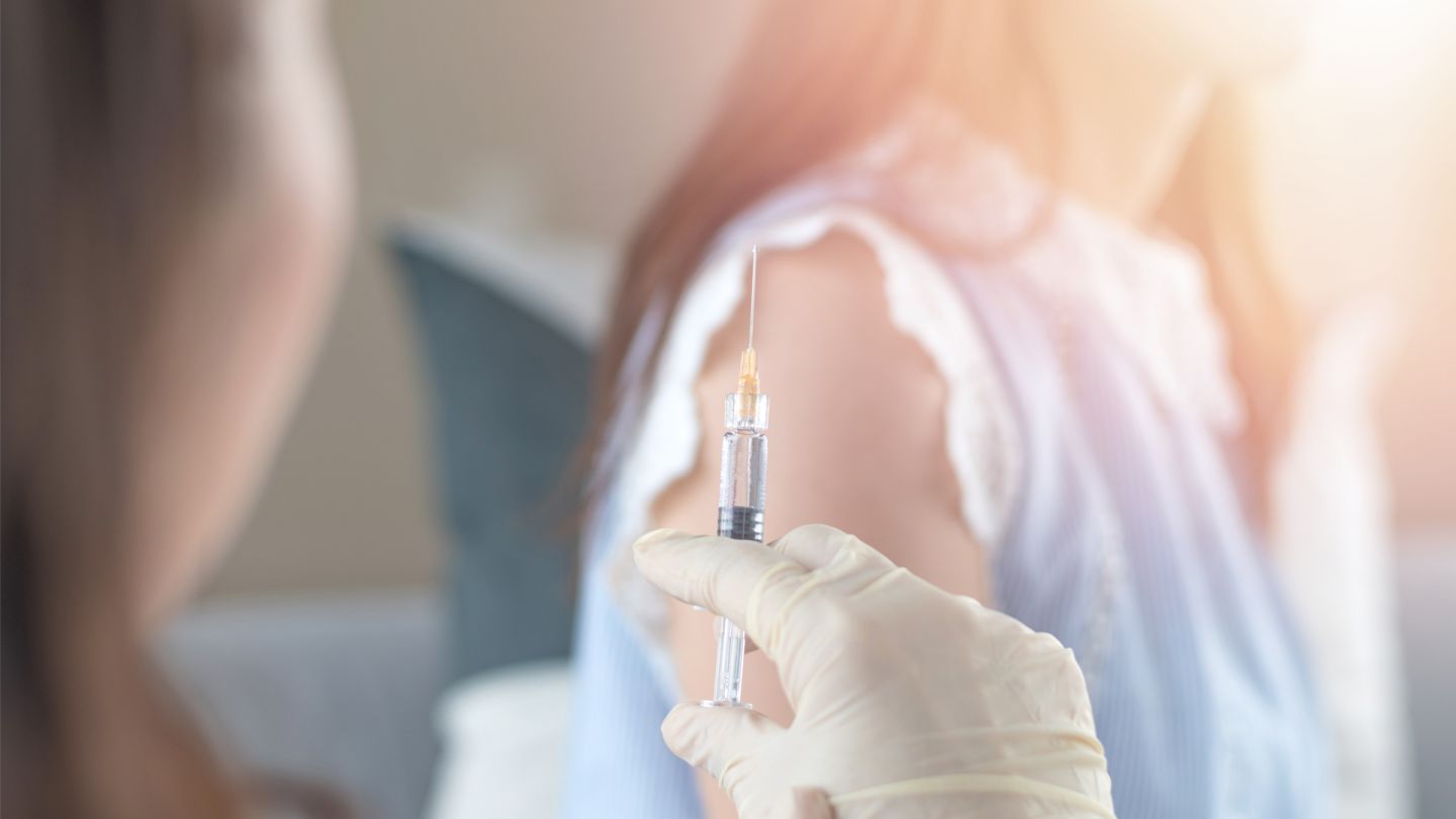 Impfungen: Eine Ärztin hält eine aufgezogene Spritze in der Hand. Im Hintergrund sitzt eine Patientin.