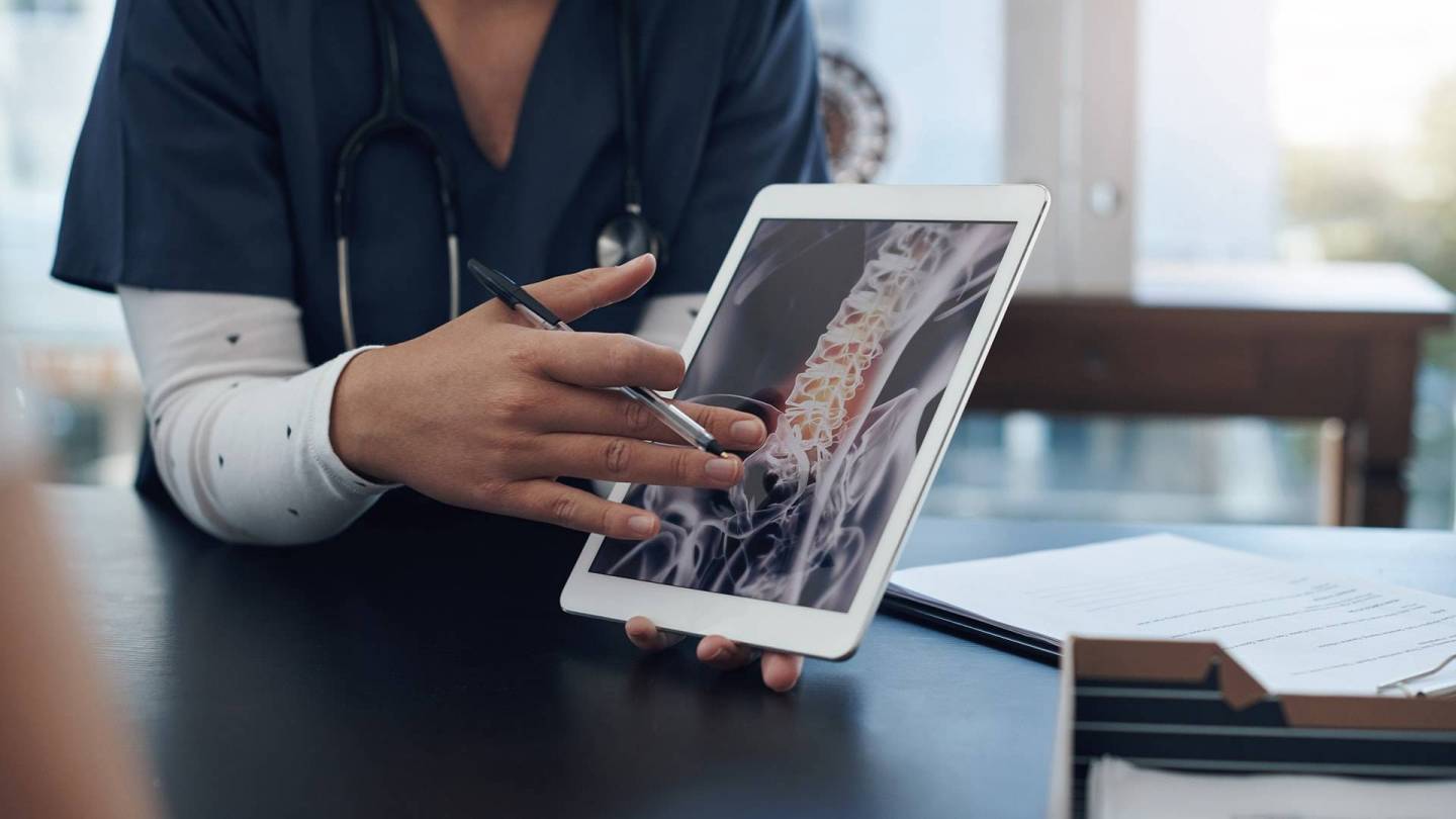 Eine Ärztin oder Pflegekraft zeigt einer anderen Person ein Bild von einer Wirbelsäule auf einem Tablet.