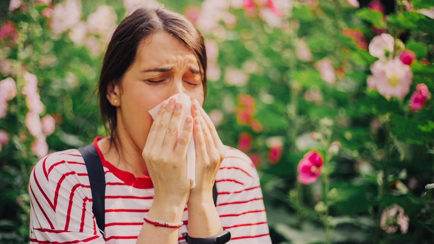 Woman in a tee shirt sneezing into a handkerchief. Flowers in bloom are visible in the background.