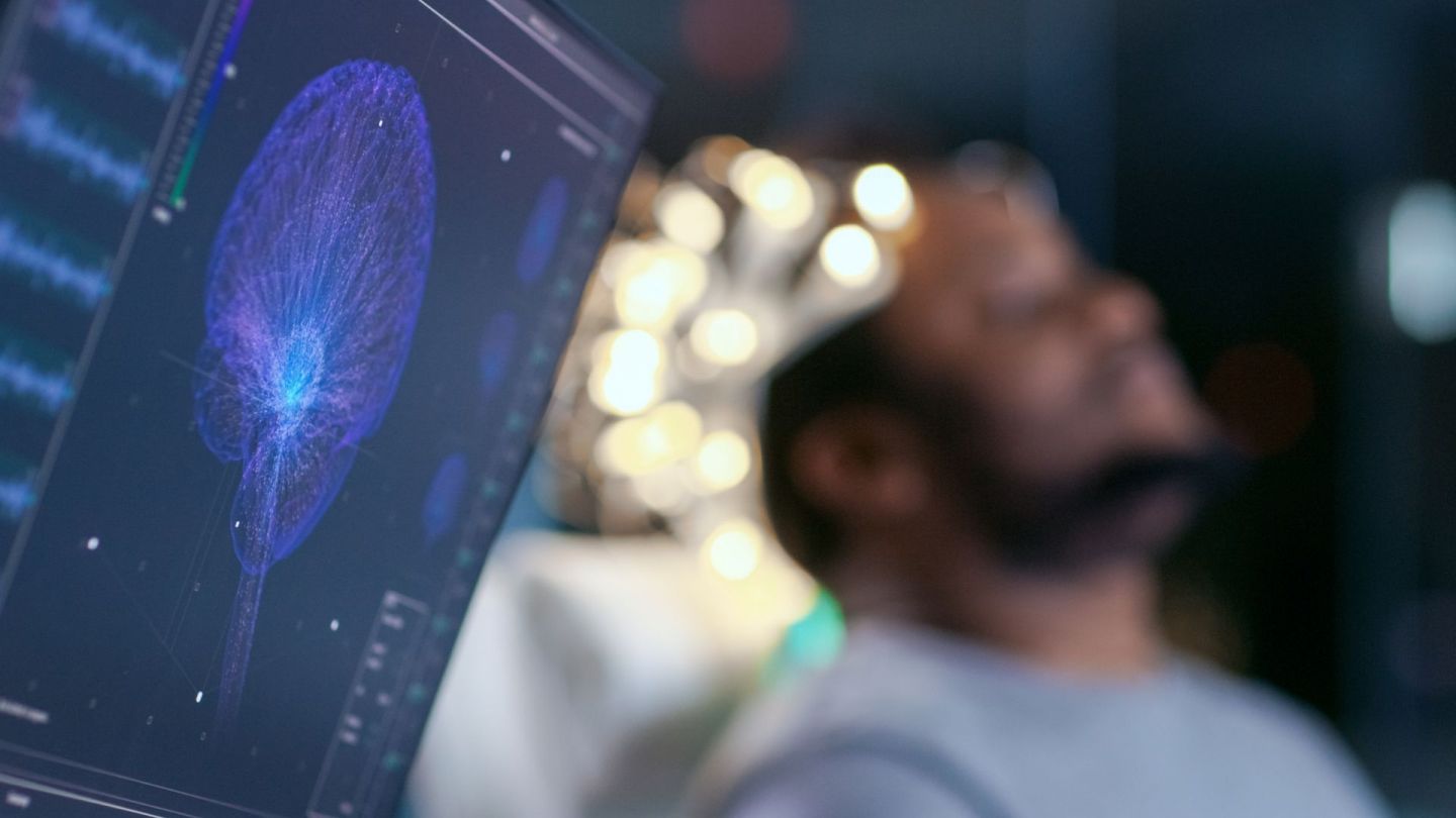 Ein EEG-Monitor zeigt das Bild eines gescanntes Schädels, durch den Nervenbahnen verlaufen. Im Hintergrund sitzt ein Mann auf einem Arztstuhl. Er hat seinen Kopf leicht nach hinten an die Lehne geneigt und scheint seine Aufen geschlossen zu haben.