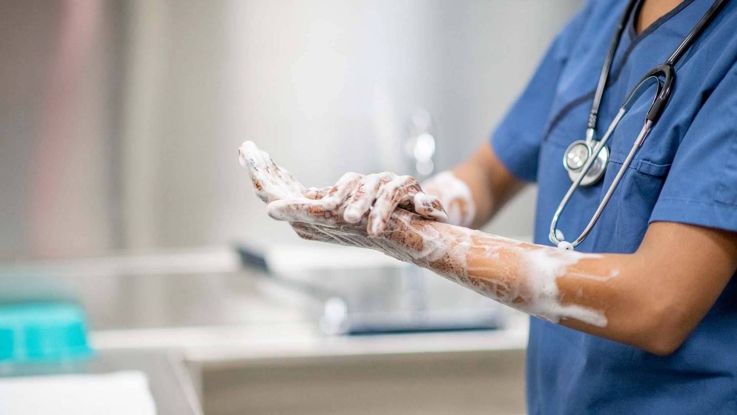 Medizinische Fachkraft in blauer OP-Kleidung wäscht sich Hände und Unterarme mit Desinfektionsseife