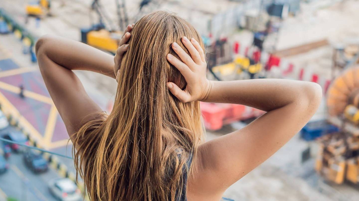 Young woman covering her ears seen from behind. Urban environment with building sites and traffic out of focus in front of her.