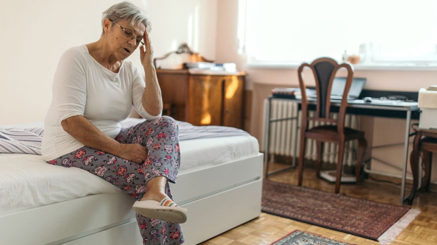 Positional vertigo: older woman sitting on a bed with her upper body leaning forward slightly, clutching her forehead with her left hand. The woman’s mouth is open slightly. She looks exhausted.