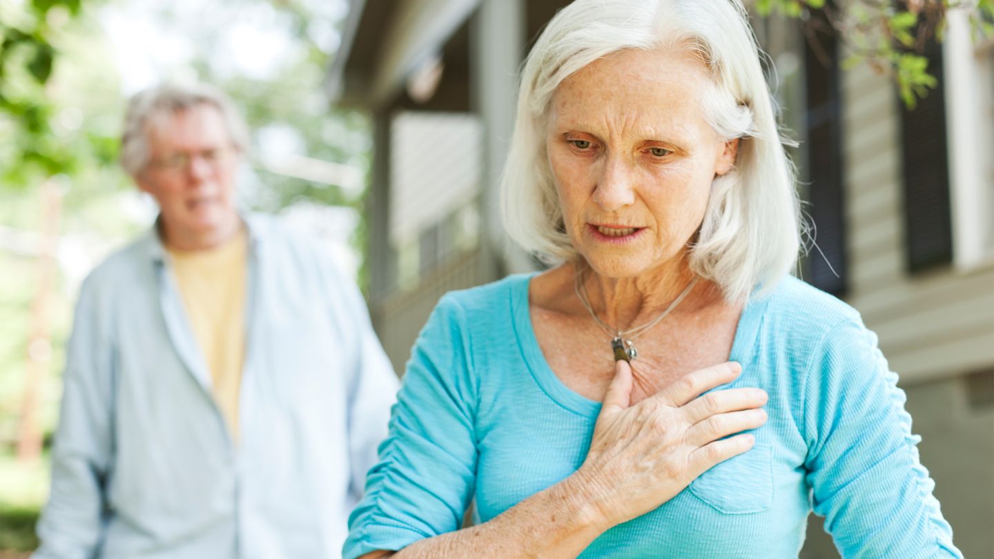 Lungenembolie: Eine ältere Frau steht im Garten und hält eine Hand an ihren Brustkorb. Im Hintergrund steht ihr Mann, beide scheinen erschrocken.