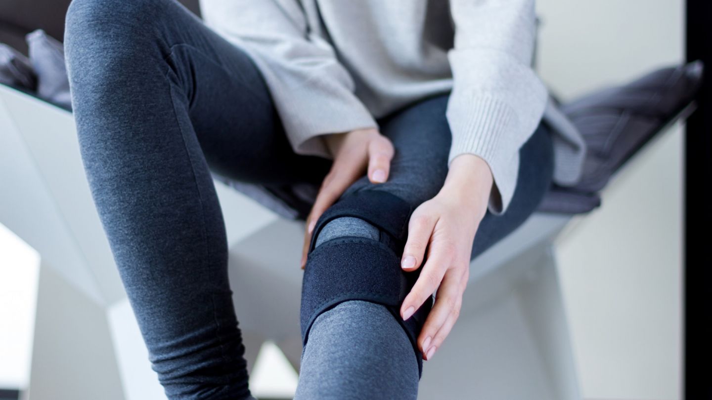 Menisküs yırtığı: Bir kadın sol bacağını uzatarak bir sandalyede oturuyor. Sol dizine bir destek bandajı bağlı.