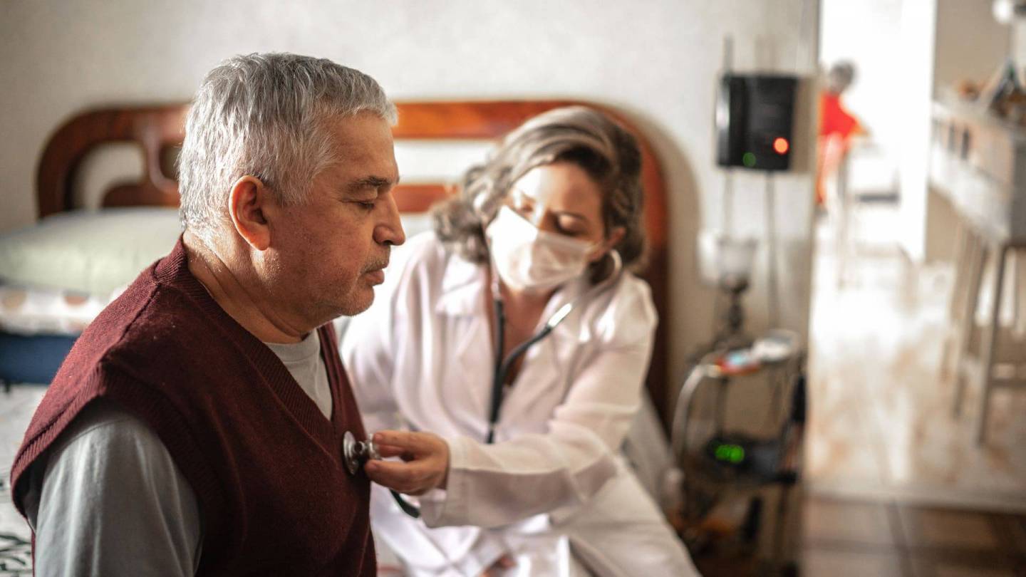 Ein älterer Mann sitzt auf einem Krankenbett. Sein Gesichtsausdruck deutet auf körperliche Schwäche hin. Eine Ärztin mit Mundschutz hört sein Herz mit dem Stethoskop ab.