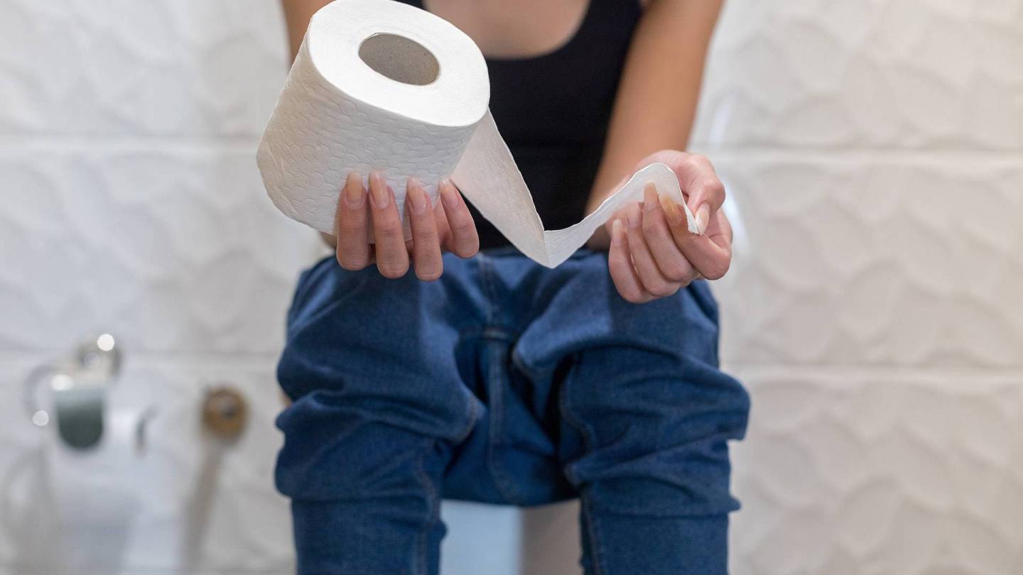 A woman sits bent forward on the toilet, holding a roll of toilet paper in her hand.