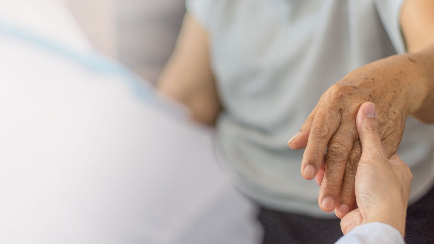 Parkinson: Eine Frau in einem weißen Kittel, offenbar eine Pflegerin, nimmt eine ältere Frau an beide Hände.