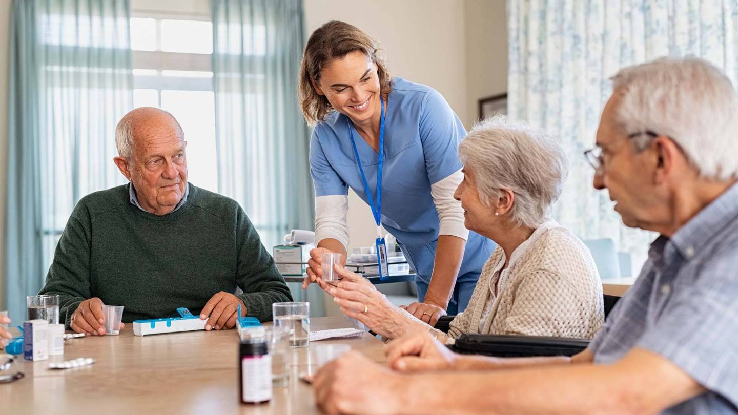 Ältere Menschen am Tisch erhalten Medikamente von einer Pflegerin.