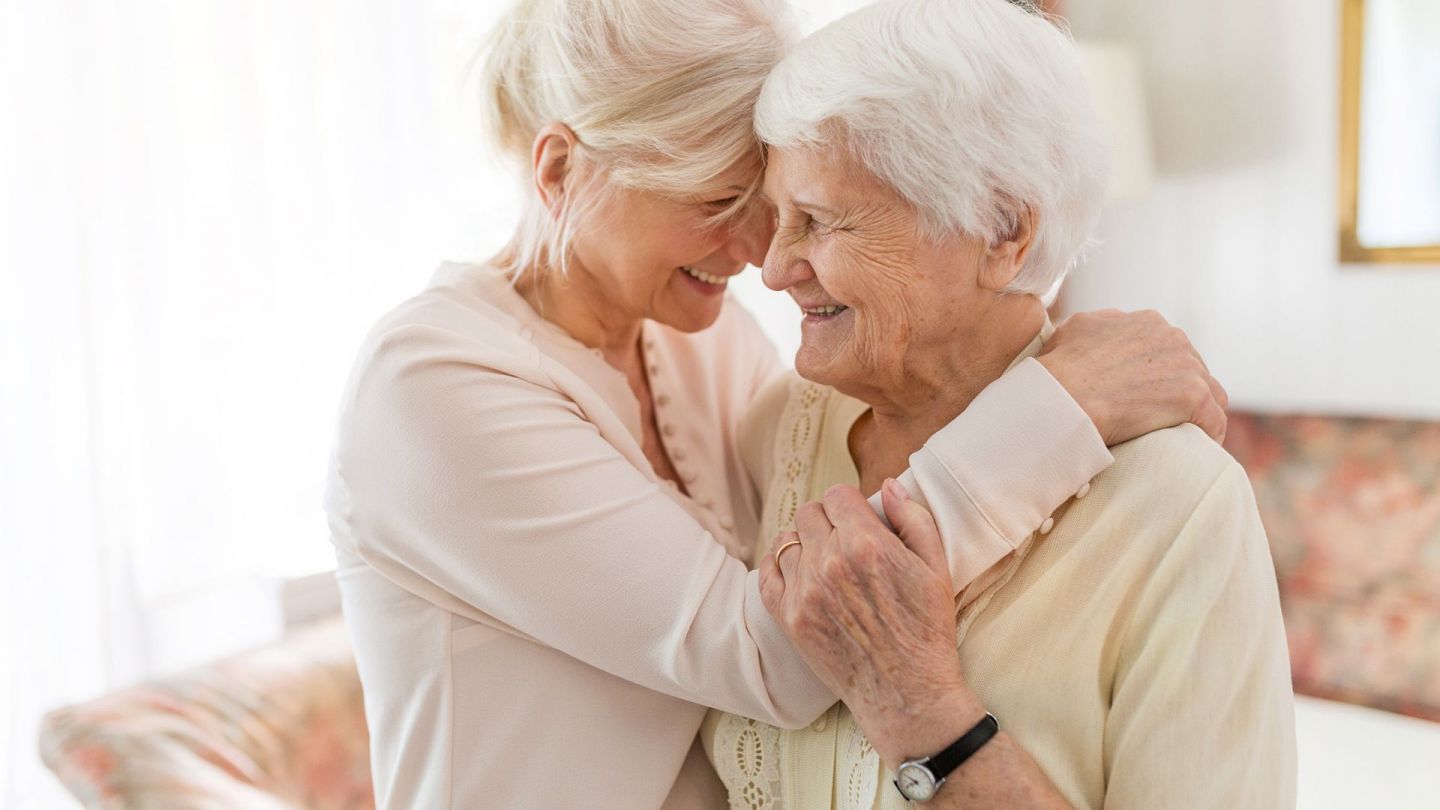 Long-term care insurance – assistance for family caregivers: mother and daughter standing opposite one another and laughing. The daughter is putting her arm around the lady’s shoulder. The lady is holding tight to the younger woman’s arm with one hand.