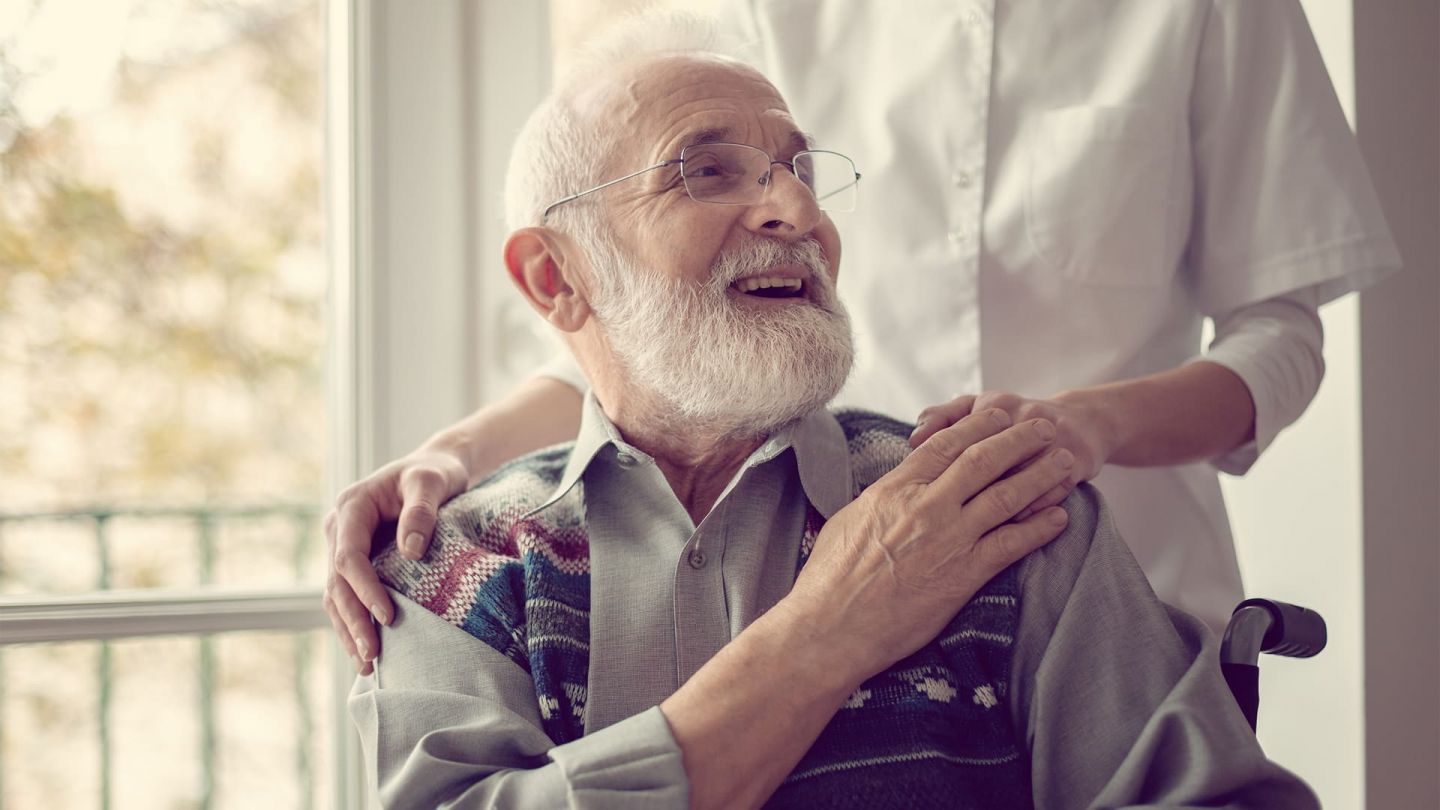 Pflegebegutachtung: Ein älterer Mann mit Bart und Brille, der im Rollstuhl sitzt, wendet sich lächelnd nach hinten zu einer Person in weiß, die hinter ihm steht und ihm die Hände auf die Schultern legt.
