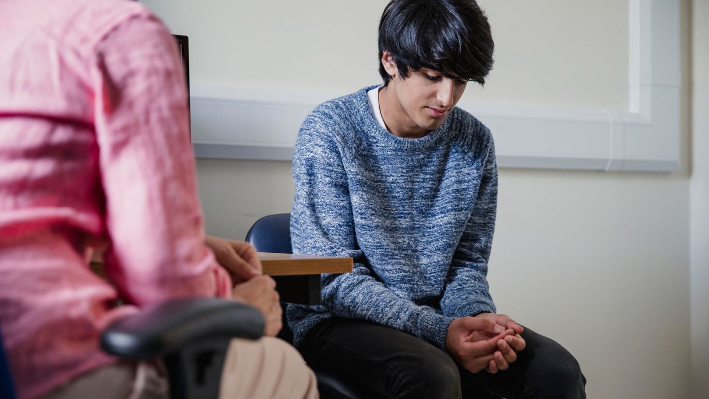 Фимоз: подросток сидит на стуле в кабинете врача. Его руки сложены на коленях, он выглядит подавленным.