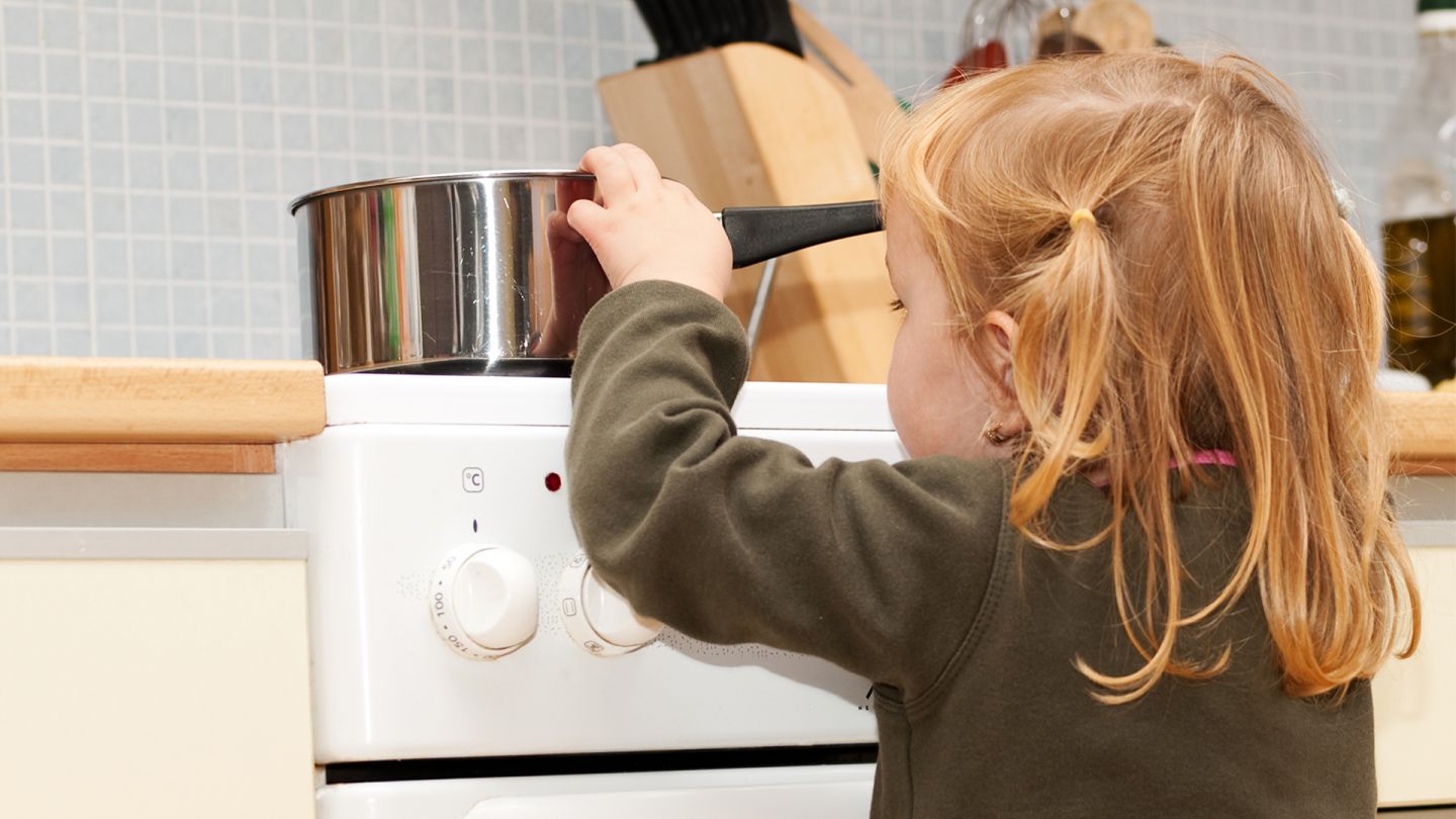 Ein kleines Mädchen steht vor einem Küchenherd auf dem ein Topf mit kochendem Inhalt steht und greift an den Topfrand, um in den Topf rein zu schauen.