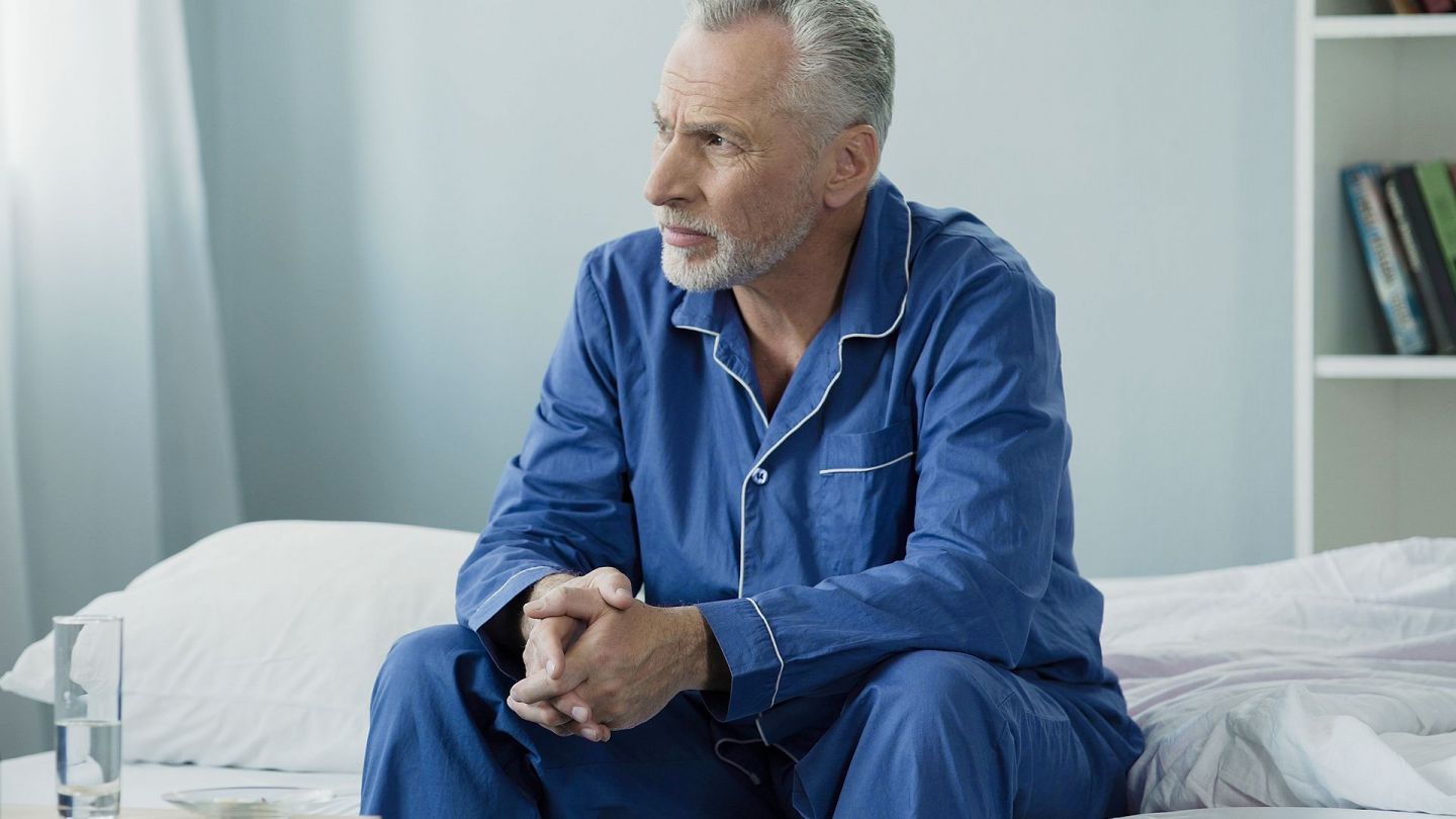 Gutartige Prostatavergrößerung (Prostatahyperplasie): Ein Mann mittleren Alters sitzt am Rand eines Krankenbettes. Er hat seine Hände ineinander gefaltet und schaut nachdenklich in Richtung Fenster.