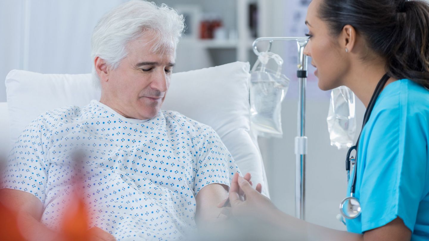 Prostate cancer: older man sitting in a hospital bed. He is looking down, a nurse is sitting opposite him. She is looking at him and taking a blood sample.