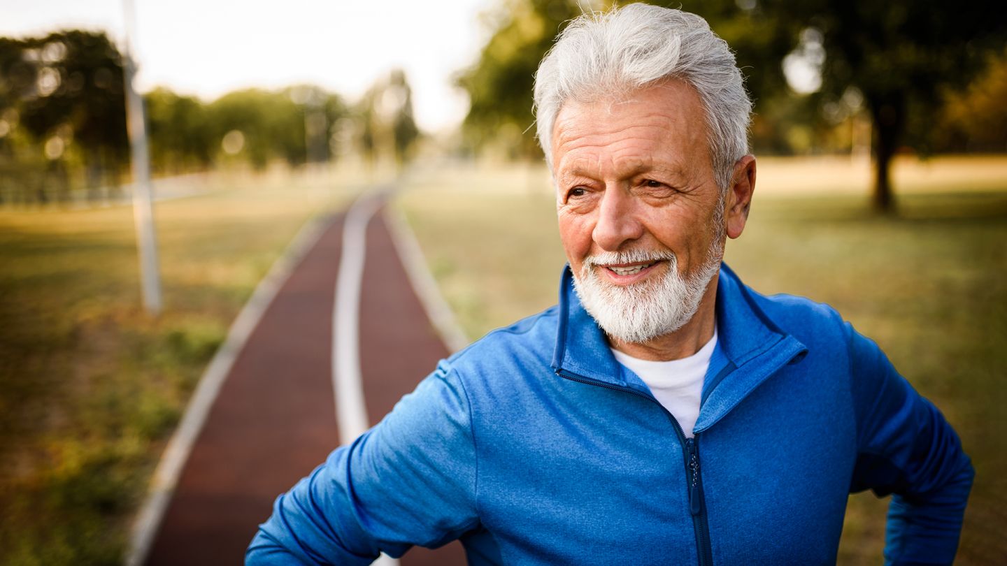 Prostatasteine: Ein älterer Herr steht in einem Park, im Hintergrund ist ein Weg zu sehen. Er trägt Sportkleidung und wirkt fit und zufrieden.