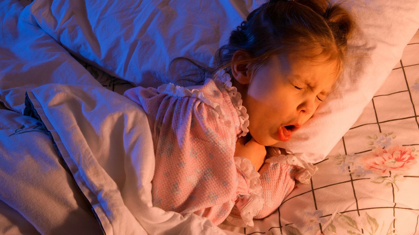 Pseudokrupp: Ein kleines Mädchen liegt in einem Bett. Sie hat sich leicht auf die Seite gedreht, den Mund weit geöffnet und hustet stark.