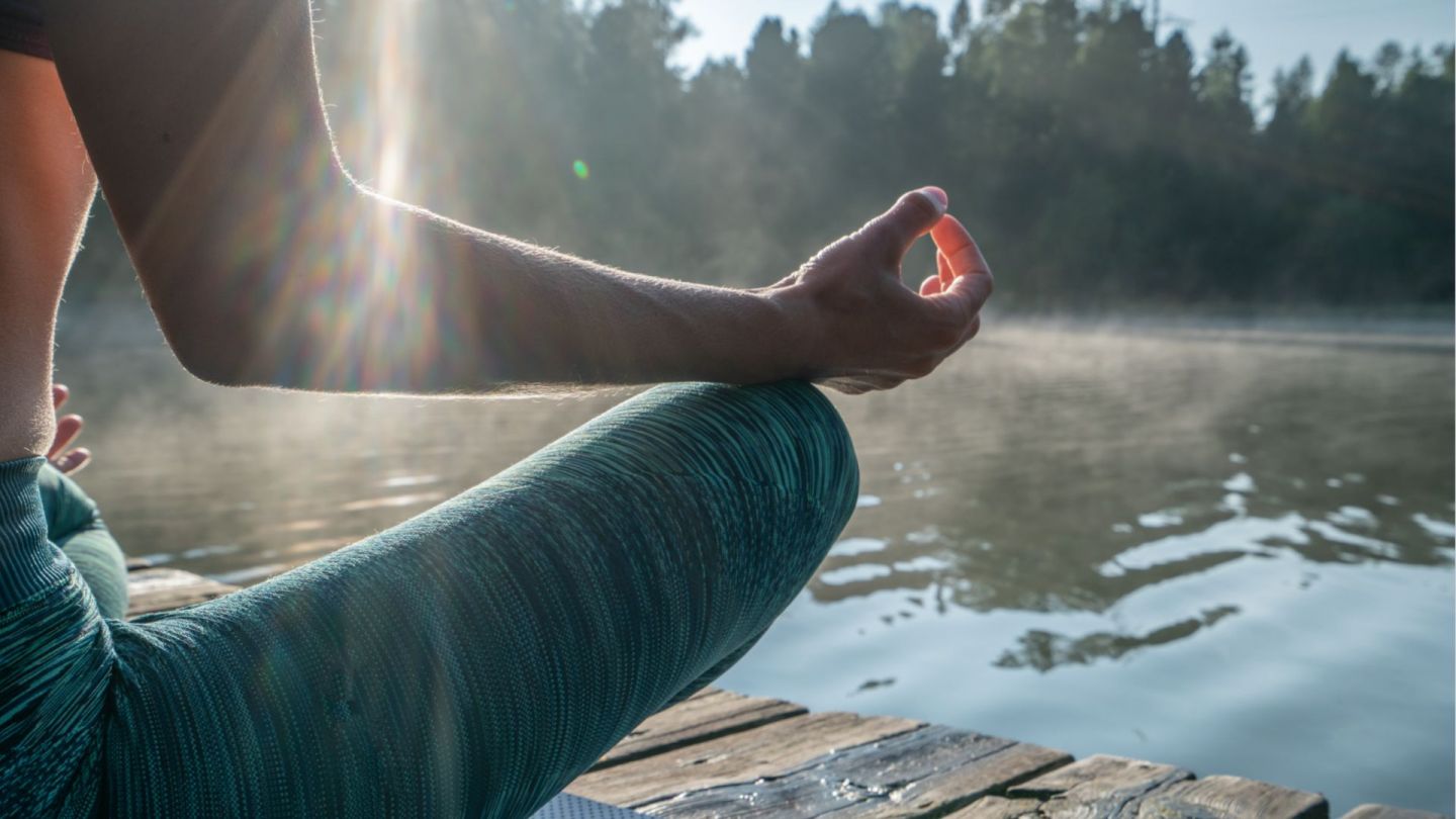 Man sitting cross-legged on a wooden jetty by a lake. The man has his right arm raised and his fingers are forming a mudra. He appears to be meditating, or possibly relaxing during yoga.