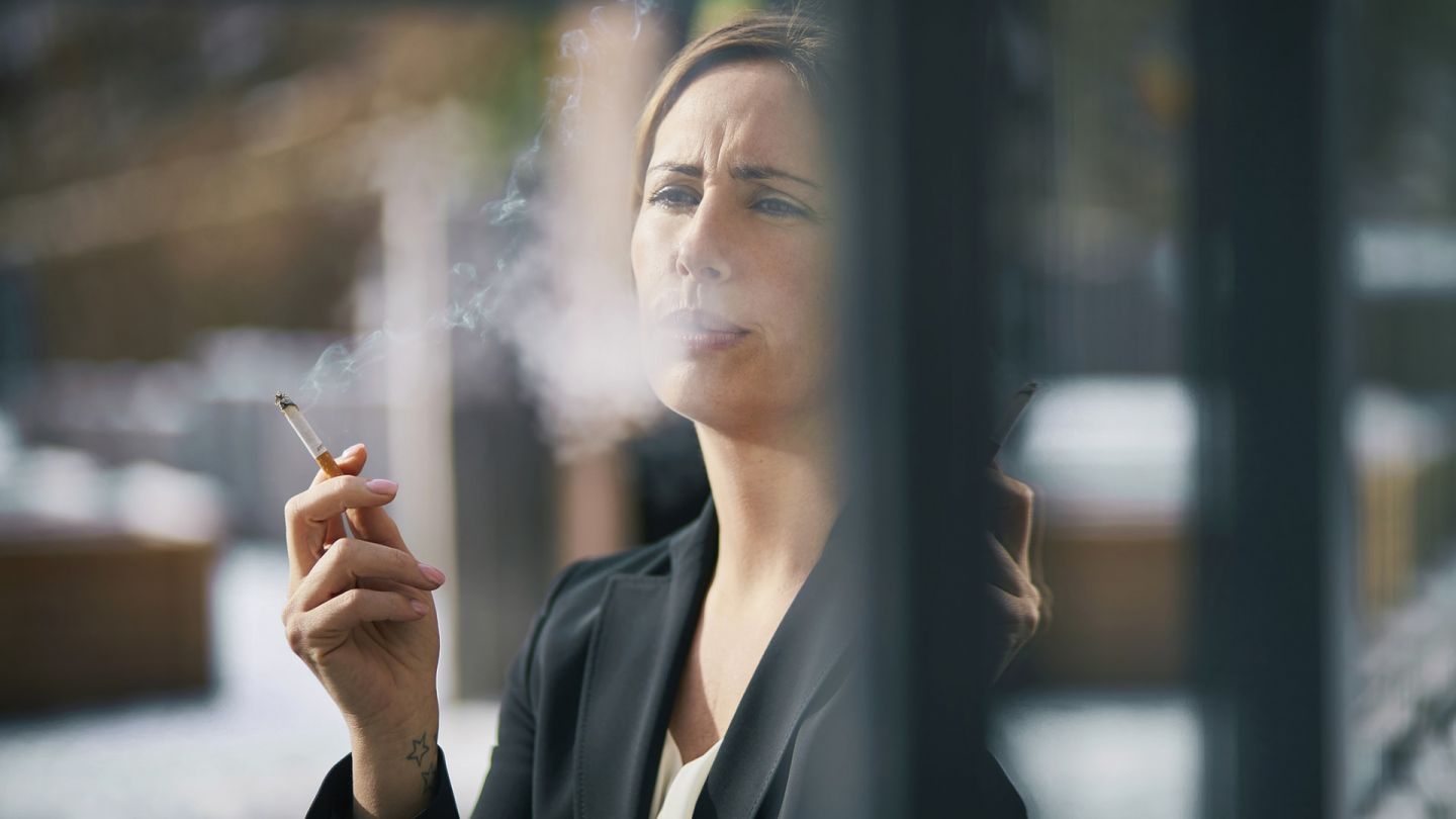 Rauchen: Eine Frau steht an einem Fenster, hält eine Zigarette in der Hand und atmet Rauch aus. Rauchschwaden umgeben ihr Gesicht.