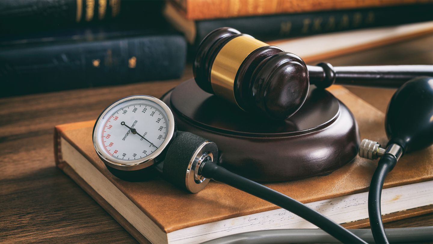 Patients’ rights: mechanical sphygmomanometer lying together with a judge’s gavel on a large book on a huge wooden table. There are books in the background.